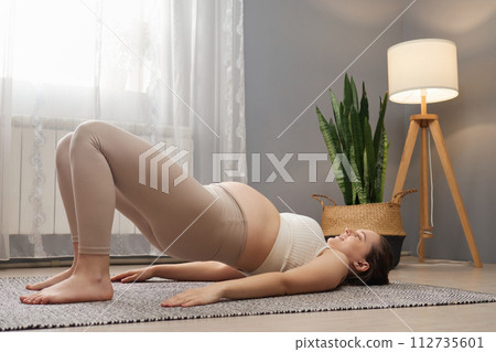 Sporty pregnant woman wearing beige top and leggings doing bridge exercise training her body taking care of health during pregnancy enjoying home gymnastic 112735601