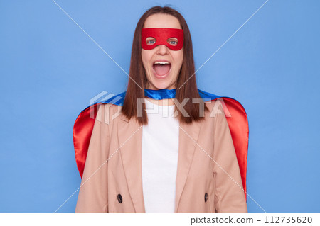 Excited amazed surprised woman wearing superhero costume standing isolated over blue background yelling with excitement rejoicing something pleasant 112735620