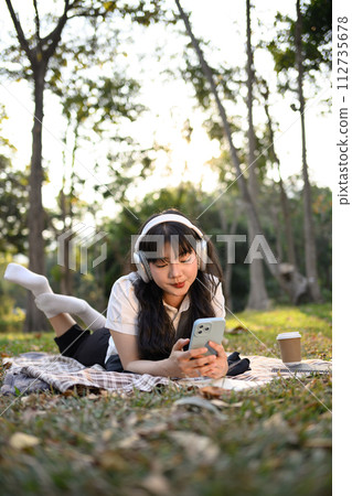 Portrait of relaxed young woman listening to music and using mobile phone on grass Portrait of relaxed young woman listening to music and using mobile phone on grass 112735678