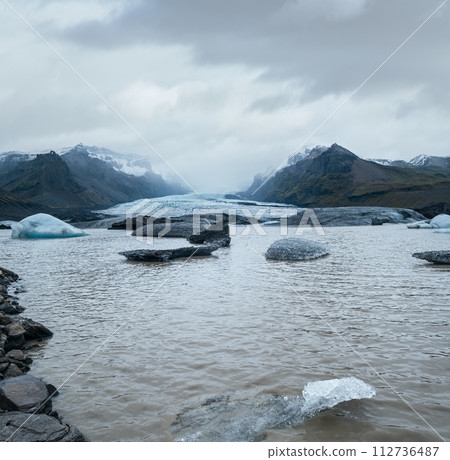 Glacier tongue slides from Vatnajokull icecap or Vatna Glacier, Iceland. Glacial lagoon with ice blocks and surrounding mountains. 112736487