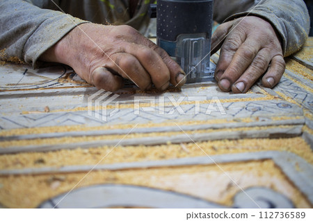 Close Up of Man Hands carving on wooden furniture. Carpenter using tool making Artistic Wood Carving. Producing an Ornament on Wood with Chisel and Hammer on a Panel of Wood . High quality photo 112736589