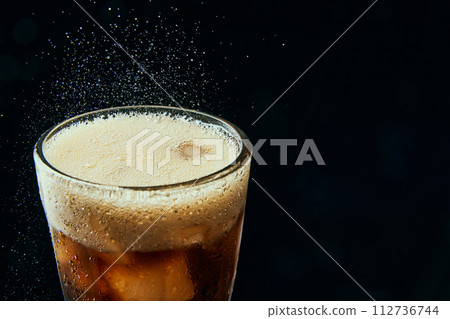 Glass wit foamy drink, soda water, coke, dark beer with ice cubes against black background. Glass wit foamy drink, soda water, coke, dark beer with ice cubes against black background. 112736744