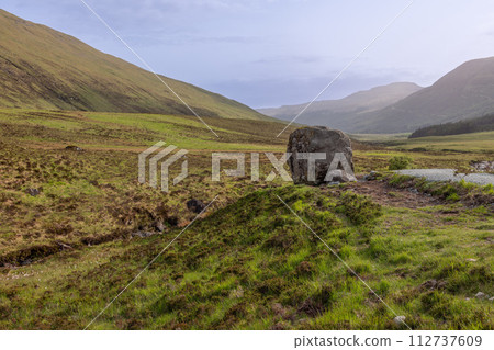 A solitary boulder stands in a sweeping Skye valley, framed by rolling hills under a soft sky A solitary boulder stands in a sweeping Skye valley, framed by rolling hills under a soft sky 112737609