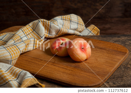 Cutting board with two saturn peaches or flat peaches on wooden background with yellow kitchen towel.. Cutting board with two saturn peaches or flat peaches on wooden background with yellow kitchen towel.. 112737612