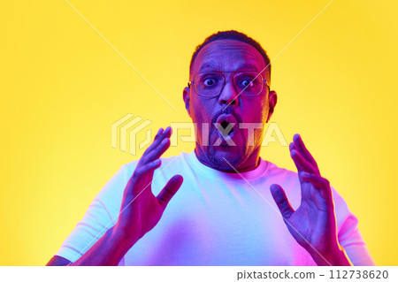 Expressive African-American man in white t-shirt showing emotions of shock against yellow studio background in neon light Expressive African-American man in white t-shirt showing emotions of shock against yellow studio background in neon light 112738620