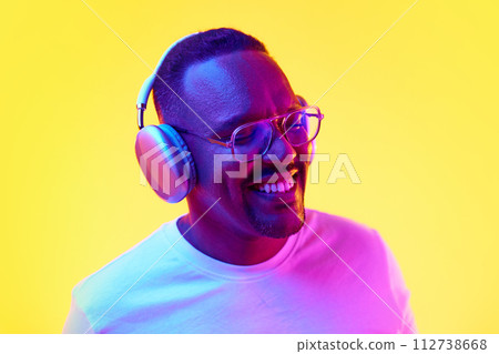 Smiling African-American man listening to music in headphones against yellow studio background in neon light Smiling African-American man listening to music in headphones against yellow studio background in neon light 112738668