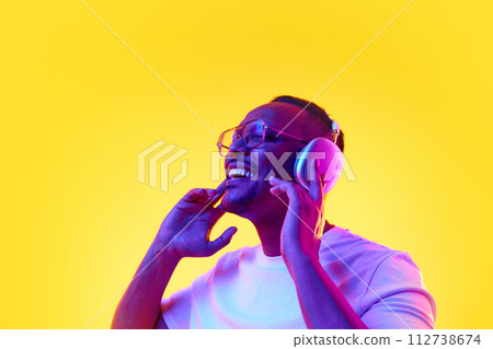 Portrait of positive African-American man listening to music in headphones and enjoying against yellow studio background in neon light Portrait of positive African-American man listening to music in headphones and enjoying against yellow studio background in neon light 112738674