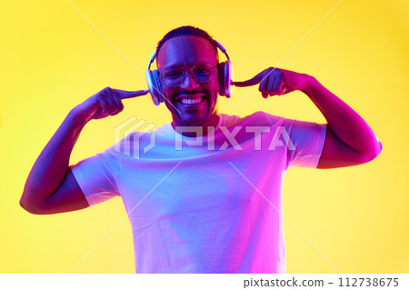 Portrait of smiling positive African-American man listening to music in headphones against yellow studio background in neon light Portrait of smiling positive African-American man listening to music in headphones against yellow studio background in neon light 112738675