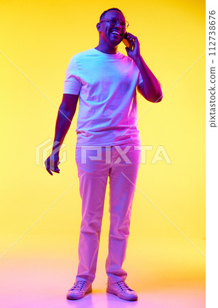 Full-length image of African-American man in casual clothes cheerfully talking on mobile phone against yellow studio background in neon light Full-length image of African-American man in casual clothes cheerfully talking on mobile phone against yellow studio background in neon light 112738676