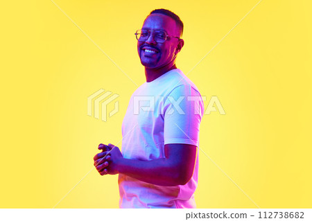Portrait of smiling African-American man in glasses and white t-shirt standing against yellow studio background in neon light 112738682