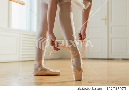 Ballerina puts on pointe shoes on leg in dance class. Classical ballet dancer woman in dance studio. Ballerina putting on tying classical pointe shoes for dance training. Ballet school concept 112738975