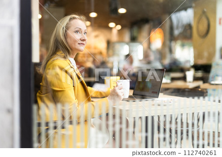 Business lunch - businesswoman sits at table in cafe by window with a laptop and drinks coffee. View through window 112740261