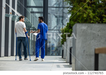Man with crutches and a male nurse in a blue uniform walking in a hospital yard 112740765
