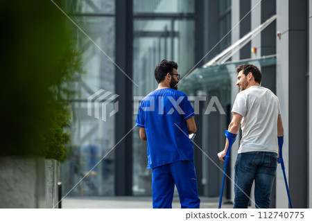 Man with crutches and a male nurse in a blue uniform walking in a hospital yard Man with crutches and a male nurse in a blue uniform walking in a hospital yard 112740775