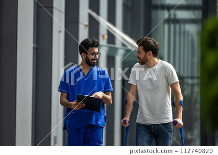 Young bearded man with crutches walking and talking to a male nurse 112740820