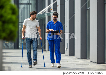 Young bearded man with crutches walking and talking to a male nurse 112740822