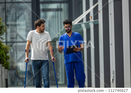Young bearded man with crutches walking and talking to a male nurse 112740827