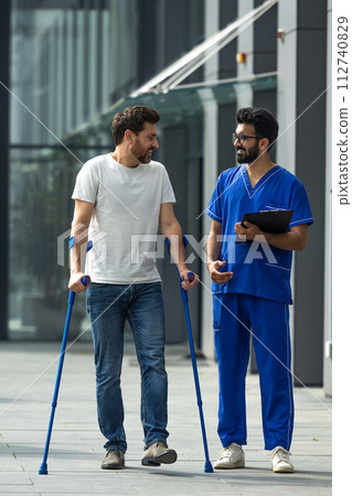 Young bearded man with crutches walking and talking to a male nurse 112740829
