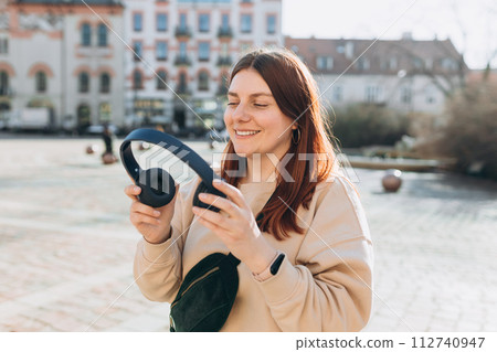 Happy young caucasian woman in good mood listening to music with headphones and standing on the street in the city. Music lover enjoying music. Urban lifestyle concept. 112740947