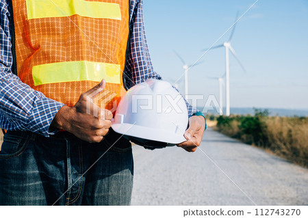 Engineer stands at windmill field holding safety helmet. Symbolizes renewable success innovation addressing global warming. Demonstrates industry leadership safety commitment. 112743270