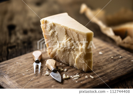 A block and pieces of Parmesan cheese on a cutting board along with a chisel and a skewer 112744408