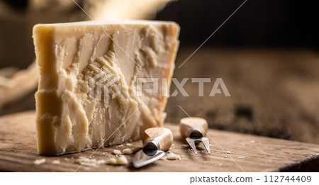 A block and pieces of Parmesan cheese on a cutting board along with a chisel and a skewer 112744409