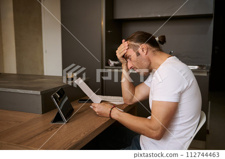 Young man sitting at the table, putting his hand on his forehead, feeling frustration and shock by costs of paying bills 112744463