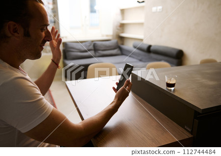 Smiling businessman sitting at desk, having video call on smart mobile phone in the office or at home. Smiling businessman sitting at desk, having video call on smart mobile phone in the office or at home. 112744484