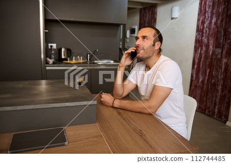 Smiling man talking on mobile phone, sitting at table at home kitchen 112744485