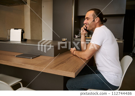 Young man smiling while talking on mobile phone, receiving call from friends, sitting at table in the home interior 112744486
