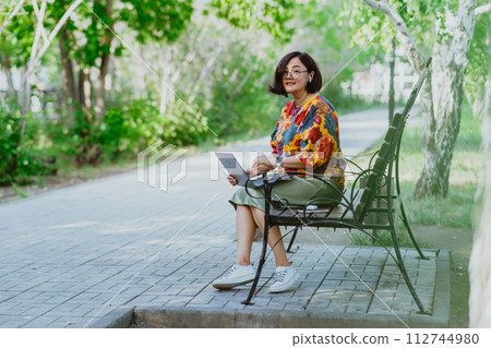 Serene outdoor office scene with a relaxed woman using her computer in nature. Female professional enjoys remote work on a calm sunny day in a lush green park 112744980