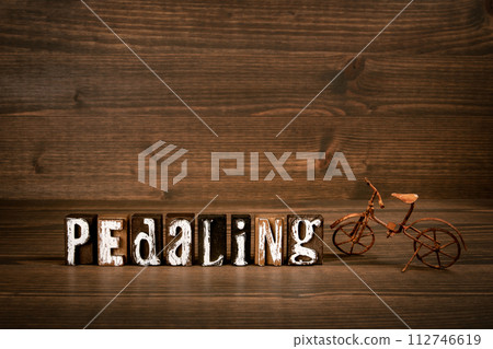 PEDALING. Text from alphabet blocks and rusty miniature bicycle on wood texture background 112746619