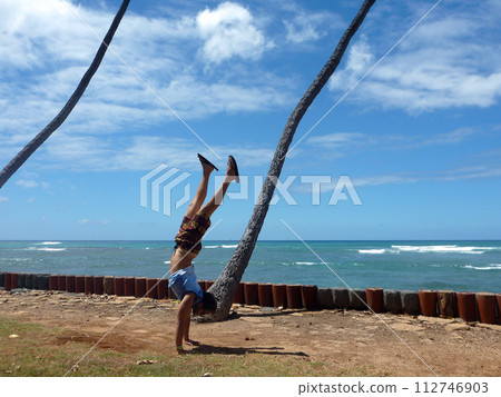 Man Handstanding along cliff shore next to shallow ocean waters of Waikiki with Coconut trees hang over Man Handstanding along cliff shore next to shallow ocean waters of Waikiki with Coconut trees hang over 112746903
