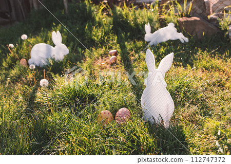 The concept of Easter holidays. Decorative Handicrafts handmade in the form of different white rabbits, located next to the painted eggs. Green grass in the background. Tint and copy The concept of Easter holidays. Decorative Handicrafts handmade in the form of different white rabbits, located next to the painted eggs. Green grass in the background. Tint and copy 112747372