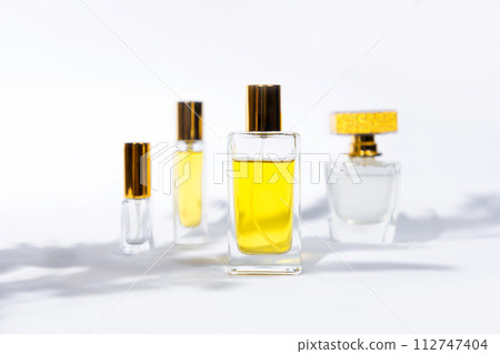 Four glass transparent bottles of golden perfume on white background with shadows of leaves. 112747404