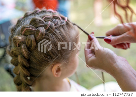 Hairdresser is braiding the hair on a young girl. Braids on the girl's head. Pink thread is woven into the hair of the pigtails. 112747727