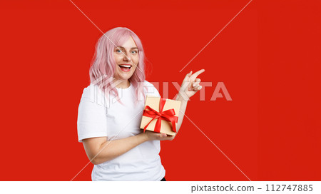 Pretty cheerful woman with pink hair holding present and showing finger away at copy space isolated over red background. Girl holding wrapped present, celebrating birthday. Special offer 112747885