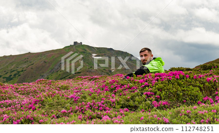 A man sits between bushes of blooming rhododendrons against the background of the mountain Pip Ivan Chornohirskyi. A man sits between bushes of blooming rhododendrons against the background of the mountain Pip Ivan Chornohirskyi. 112748251