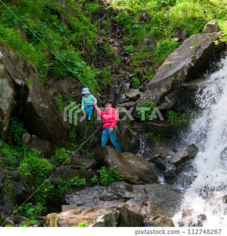 Mother and daughter are standing near a mountain waterfall. Mother and daughter are standing near a mountain waterfall. 112748267