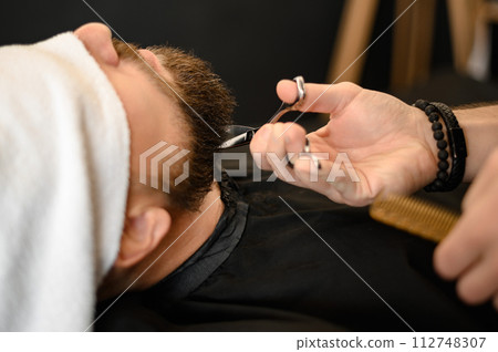 A barber stylist trims the beard of a Caucasian man, whose face is covered with towel, with scissors 112748307