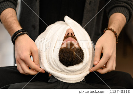 The barber covers the client face with a hot wet towel to steam off the beard. The barber covers the client face with a hot wet towel to steam off the beard. 112748318