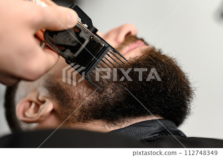 Cutting a gentlemans beard in a barbershop with a clipper. Shortening the length of the beard from the sides by the master for the client. 112748349