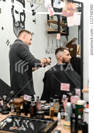 A barber cuts a man with a beard in a barber shop. Short haircut of the client with clipper. 112748360