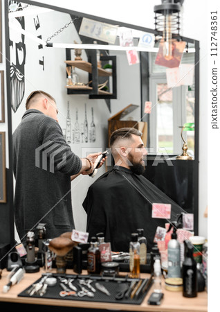 A barber cuts a man with a beard in a barber shop. Short haircut of the client with clipper. A barber cuts a man with a beard in a barber shop. Short haircut of the client with clipper. 112748361