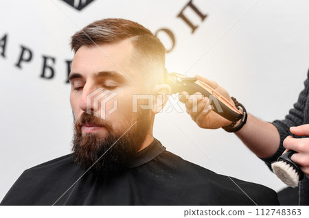 A barber cuts a man with a beard in a barber shop. Short haircut of the client with clipper. A barber cuts a man with a beard in a barber shop. Short haircut of the client with clipper. 112748363
