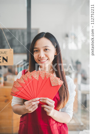 A bright-eyed young woman in an apron presents traditional red envelopes, her smile reflecting the cultural joy and hospitality associated with this gesture. A bright-eyed young woman in an apron presents traditional red envelopes, her smile reflecting the cultural joy and hospitality associated with this gesture. 112752211