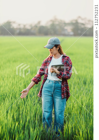 Diligent female agronomist uses a tablet to assess crop health in a lush green field during early evening. 112752231