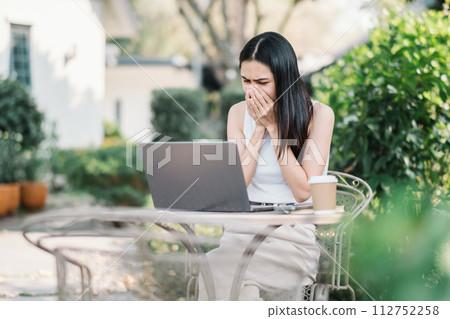 Worried young woman covering her mouth, looking at her laptop screen with concern, seated at a garden table with a coffee cup. 112752258