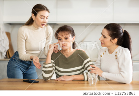 Sad woman sits silently in kitchen, adult girls sympathize and comfort her Sad woman sits silently in kitchen, adult girls sympathize and comfort her 112755865