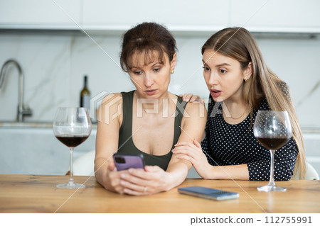 Two sad middle-aged women looking at the phone display while sitting at the table at home Two sad middle-aged women looking at the phone display while sitting at the table at home 112755991